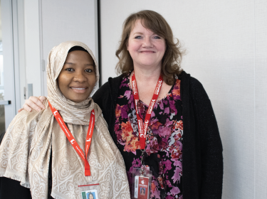 Woman wearing a hijab smiling at the camera next to another woman smiling att he camera